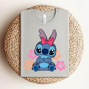 Puede incluir: Una camiseta gris claro con un personaje de dibujos animados azul, con un lazo rosa y ojos grandes. Flores de hibisco rosas y naranjas rodean al personaje. La camiseta est&aacute; doblada sobre una superficie circular tejida.