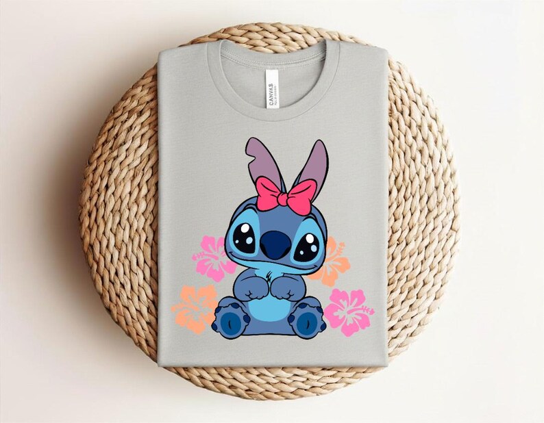Puede incluir: Una camiseta gris claro con un personaje de dibujos animados azul, con un lazo rosa y ojos grandes. Flores de hibisco rosas y naranjas rodean al personaje. La camiseta est&aacute; doblada sobre una superficie circular tejida.