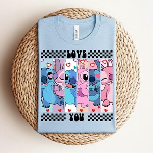 May include: Light blue t-shirt featuring a graphic of Stitch and Angel characters with the words "LOVE YOU". The design includes hearts and a checkered pattern, perfect for fans of the animated series.