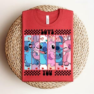 May include: Red t-shirt featuring a graphic of cartoon characters in shades of blue and pink, with the words "LOVE YOU" in black. The design includes hearts and a checkered pattern. The shirt is displayed on a woven, circular surface.