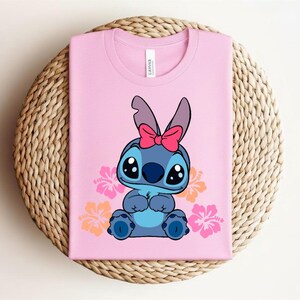 Puede incluir: Camiseta rosa con un personaje de dibujos animados de pelaje azul, ojos grandes y un lazo rosa. El dise&ntilde;o incluye flores naranjas y rosas. La camiseta est&aacute; doblada sobre una estera redonda tejida.