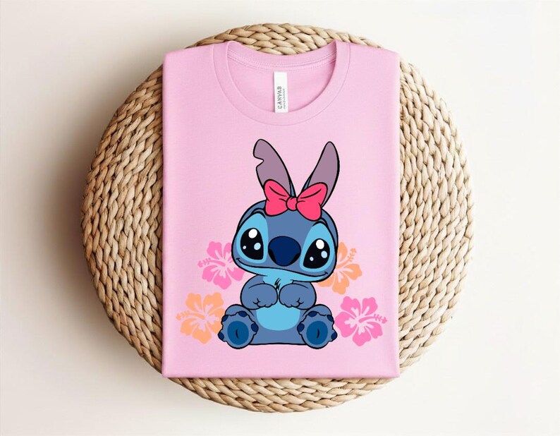 Puede incluir: Camiseta rosa con un personaje de dibujos animados de pelaje azul, ojos grandes y un lazo rosa. El dise&ntilde;o incluye flores naranjas y rosas. La camiseta est&aacute; doblada sobre una estera redonda tejida.
