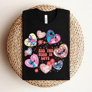 Peut inclure: T-shirt noir avec des personnages de dessins anim&eacute;s en forme de c&oelig;ur et le texte "All You Need Is Love". Les c&oelig;urs sont roses, bleus et jaunes, avec des motifs floraux. Le t-shirt est pr&eacute;sent&eacute; sur une surface tress&eacute;e.