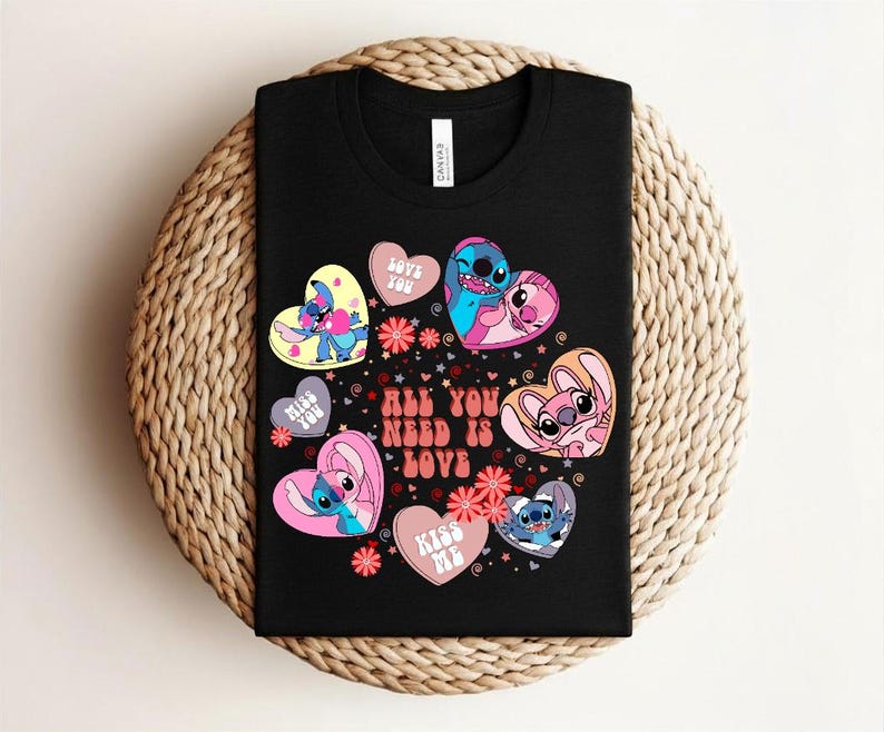 Peut inclure: T-shirt noir avec des personnages de dessins anim&eacute;s en forme de c&oelig;ur et le texte "All You Need Is Love". Les c&oelig;urs sont roses, bleus et jaunes, avec des motifs floraux. Le t-shirt est pr&eacute;sent&eacute; sur une surface tress&eacute;e.