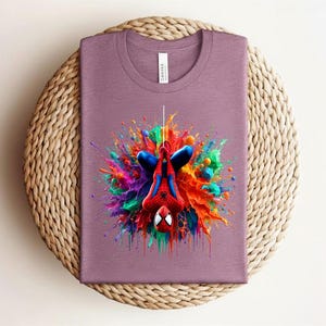 May include: A mauve t-shirt featuring a colorful graphic of Spider-Man hanging upside down. The design includes vibrant splashes of red, blue, green, and yellow, creating a dynamic and eye-catching effect. The shirt is folded on a woven surface.