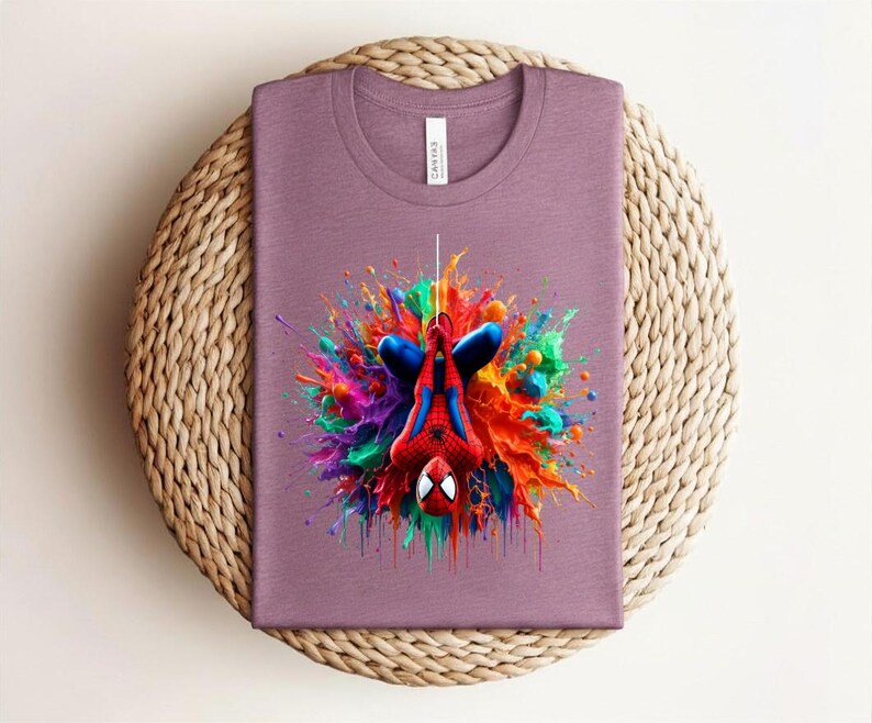 May include: A mauve t-shirt featuring a colorful graphic of Spider-Man hanging upside down. The design includes vibrant splashes of red, blue, green, and yellow, creating a dynamic and eye-catching effect. The shirt is folded on a woven surface.