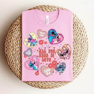 Peut inclure: Un t-shirt rose avec des personnages de dessins anim&eacute;s en forme de c&oelig;ur. Le t-shirt affiche le texte "All You Need Is Love" et d'autres phrases comme "Love You" et "Kiss Me". Le motif comprend des motifs floraux et des &eacute;toiles.