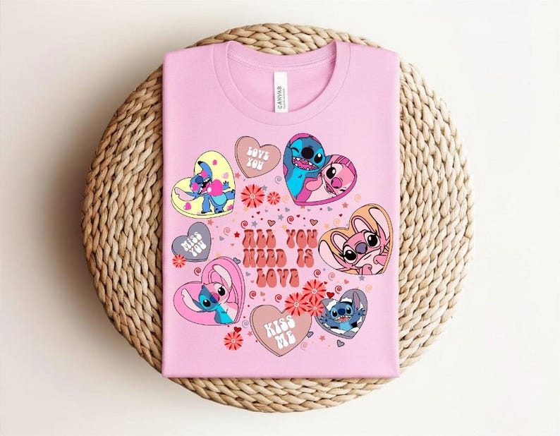 Peut inclure: Un t-shirt rose avec des personnages de dessins anim&eacute;s en forme de c&oelig;ur. Le t-shirt affiche le texte "All You Need Is Love" et d'autres phrases comme "Love You" et "Kiss Me". Le motif comprend des motifs floraux et des &eacute;toiles.