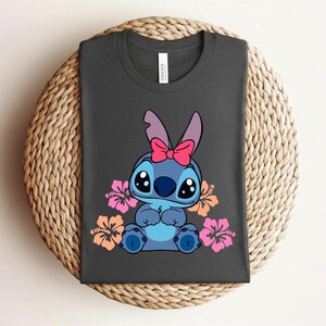 Puede incluir: Camiseta gris oscuro con un personaje de dibujos animados azul, con un lazo rosa y rodeado de flores de hibisco rosas y naranjas. La camiseta est&aacute; doblada sobre una superficie circular tejida.