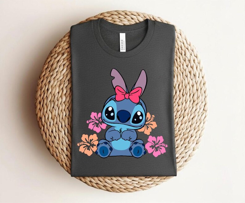 Puede incluir: Camiseta gris oscuro con un personaje de dibujos animados azul, con un lazo rosa y rodeado de flores de hibisco rosas y naranjas. La camiseta est&aacute; doblada sobre una superficie circular tejida.