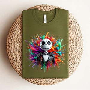 May include: An olive green t-shirt featuring a graphic of a white skull character with large black eyes and a stitched mouth, wearing a black pinstripe suit and bow tie, set against a vibrant, multi-colored paint splatter background.