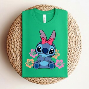 Puede incluir: Una camiseta verde con un personaje de dibujos animados de piel azul, ojos grandes y un lazo rosa. Flores de hibisco rosas y naranjas rodean al personaje. La camiseta est&aacute; doblada sobre una superficie tejida.
