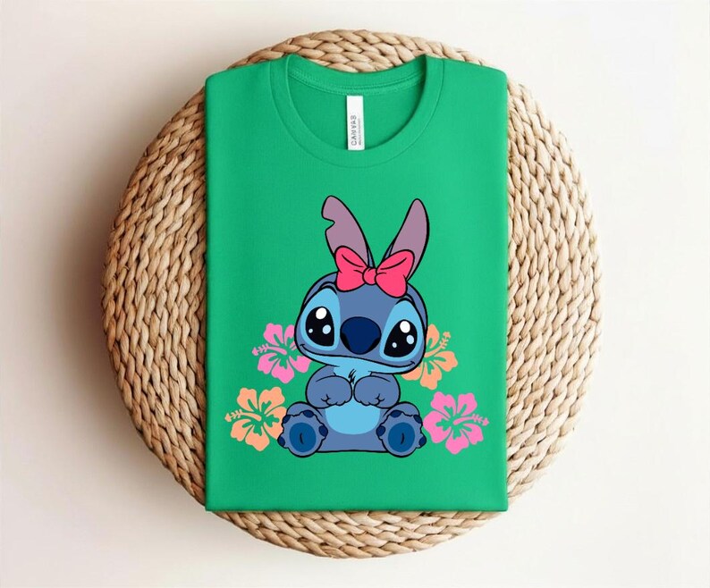 Puede incluir: Una camiseta verde con un personaje de dibujos animados de piel azul, ojos grandes y un lazo rosa. Flores de hibisco rosas y naranjas rodean al personaje. La camiseta est&aacute; doblada sobre una superficie tejida.