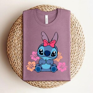 Puede incluir: Camiseta malva con un personaje de dibujos animados con ojos grandes, pelaje azul y un lazo rosa. El personaje est&aacute; rodeado de flores de hibisco rosas y naranjas. La camiseta est&aacute; doblada sobre una superficie circular tejida.
