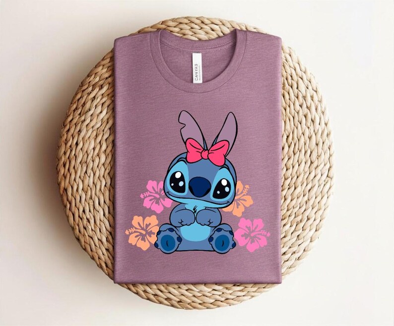 Puede incluir: Camiseta malva con un personaje de dibujos animados con ojos grandes, pelaje azul y un lazo rosa. El personaje est&aacute; rodeado de flores de hibisco rosas y naranjas. La camiseta est&aacute; doblada sobre una superficie circular tejida.