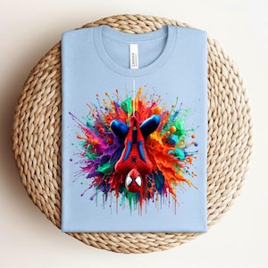 May include: Light blue t-shirt featuring a colorful graphic of Spider-Man with a vibrant paint splash design. The superhero is depicted upside down, with a red and blue suit. The shirt is folded on a woven, circular surface.