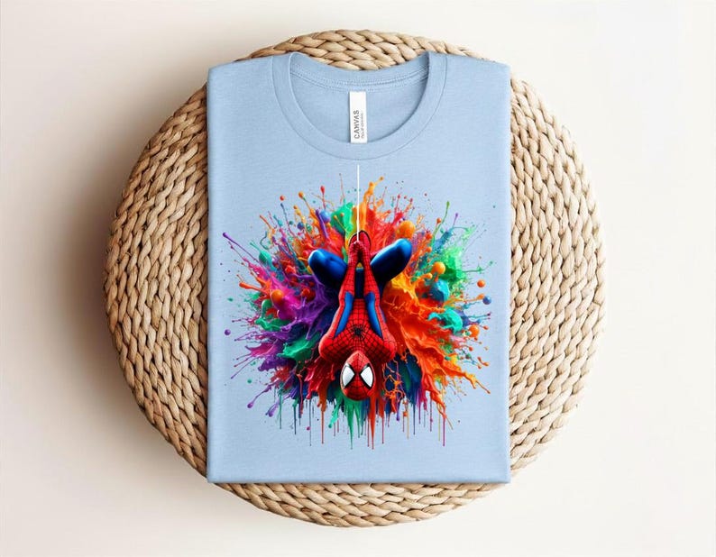 May include: Light blue t-shirt featuring a colorful graphic of Spider-Man with a vibrant paint splash design. The superhero is depicted upside down, with a red and blue suit. The shirt is folded on a woven, circular surface.