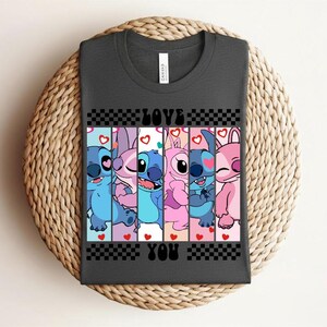 May include: Dark gray t-shirt featuring a colorful graphic of cartoon characters with the text "LOVE YOU" in black. The design includes blue, pink, and purple characters with hearts and a checkered pattern.