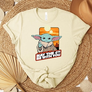 Może przedstawiać: Kremowa koszulka z grafiką Grogu z Gwiezdnych Wojen, z napisem "Star Wars Day May The 4th Be With You". Projekt zawiera pomarańczowo-ż&oacute;łte tło zachodu słońca. Koszulka jest zawiązana na dole.