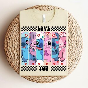 May include: A beige t-shirt featuring a colorful graphic of cartoon characters with the words "LOVE YOU" in black. The design includes blue, pink, and red hearts, and a checkered pattern. The shirt is displayed on a woven, circular surface.