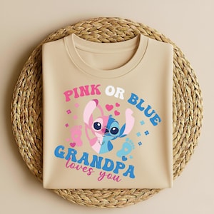 Pu&ograve; includere: T-shirt beige con un personaggio dei cartoni animati blu e rosa. Il testo "PINK OR BLUE" e "GRANDPA loves you" &egrave; in rosa e blu. La maglietta &egrave; su un tappetino rotondo intrecciato.
