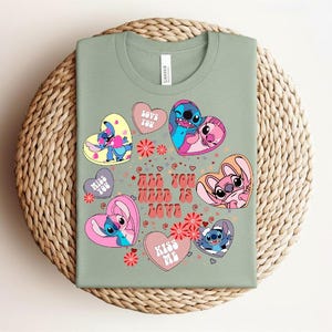 Peut inclure: T-shirt vert sauge avec des personnages de dessins anim&eacute;s dans des motifs en forme de c&oelig;ur. Les c&oelig;urs portent des phrases comme "Love You" et "Kiss Me". Le texte "All You Need Is Love" est &eacute;galement pr&eacute;sent. Le t-shirt est pr&eacute;sent&eacute; sur un tapis rond tiss&eacute;.