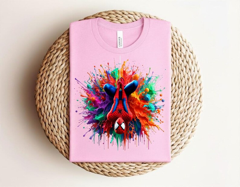 May include: A light pink t-shirt featuring a colorful graphic of Spider-Man in a dynamic pose. The superhero is depicted upside down against a vibrant splash of paint in various colors. The shirt is folded neatly on a woven surface.