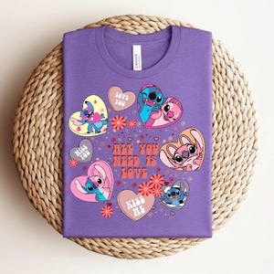 Peut inclure: Un t-shirt violet avec un motif color&eacute; de personnages de dessins anim&eacute;s en forme de c&oelig;ur. Le t-shirt porte l'inscription "All You Need Is Love" et d'autres phrases comme "Love You" et "Kiss Me".