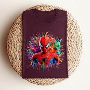 Op de afbeelding: Een bordeauxrood t-shirt met een kleurrijke afbeelding van Spider-Man. Het hoofd en de schouders van de superheld worden afgebeeld met een levendig, veelkleurig spatteffect op de achtergrond. Het shirt is op een geweven, cirkelvormig oppervlak gevouwen.