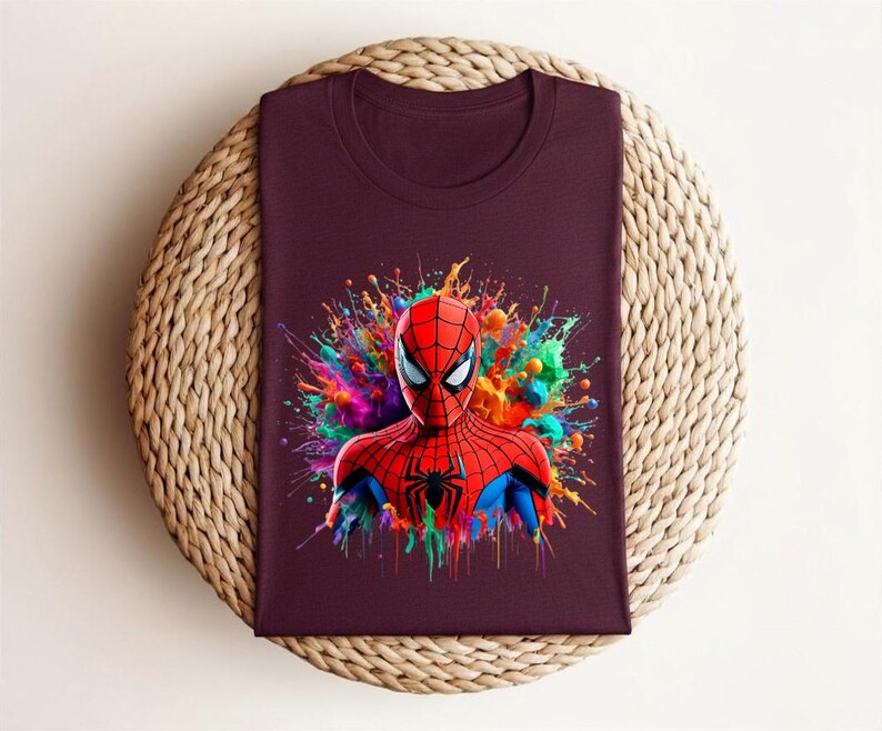 Op de afbeelding: Een bordeauxrood t-shirt met een kleurrijke afbeelding van Spider-Man. Het hoofd en de schouders van de superheld worden afgebeeld met een levendig, veelkleurig spatteffect op de achtergrond. Het shirt is op een geweven, cirkelvormig oppervlak gevouwen.