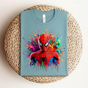 Op de afbeelding: Lichtblauw T-shirt met een afbeelding van Spider-Man. De superheld draagt een rood en zwart pak, met een kleurrijke splash achter hem. Het shirt wordt gepresenteerd op een geweven, cirkelvormig oppervlak.