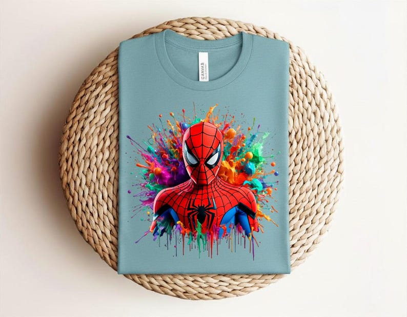 Op de afbeelding: Lichtblauw T-shirt met een afbeelding van Spider-Man. De superheld draagt een rood en zwart pak, met een kleurrijke splash achter hem. Het shirt wordt gepresenteerd op een geweven, cirkelvormig oppervlak.