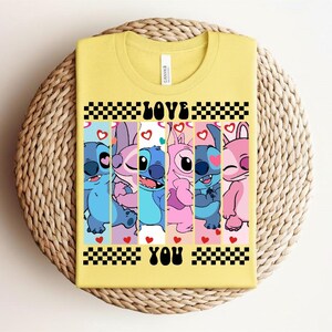 May include: Yellow t-shirt featuring a graphic of cartoon characters in blue, pink, and purple, with the words "LOVE YOU" in black. The design includes hearts and a checkered pattern.