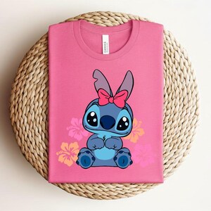 Puede incluir: Camiseta rosa con un personaje de dibujos animados con pelaje azul, ojos grandes y un lazo rosa. El dise&ntilde;o incluye flores de hibisco. La camiseta est&aacute; doblada sobre una superficie tejida.