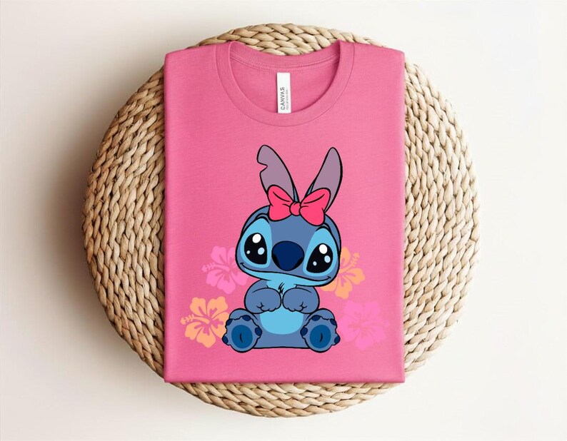Puede incluir: Camiseta rosa con un personaje de dibujos animados con pelaje azul, ojos grandes y un lazo rosa. El dise&ntilde;o incluye flores de hibisco. La camiseta est&aacute; doblada sobre una superficie tejida.