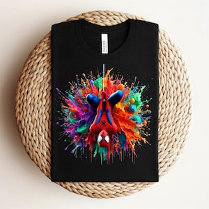 May include: Black t-shirt featuring a colorful graphic of Spider-Man hanging upside down against a vibrant splash of paint. The design includes red, blue, green, and yellow hues. The shirt is folded on a woven surface.