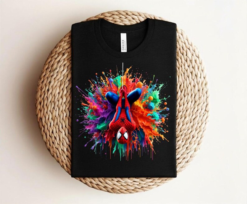 May include: Black t-shirt featuring a colorful graphic of Spider-Man hanging upside down against a vibrant splash of paint. The design includes red, blue, green, and yellow hues. The shirt is folded on a woven surface.