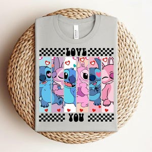 May include: A light gray t-shirt featuring a colorful graphic of cartoon characters. The design includes the words "LOVE YOU" in a checkered pattern. The characters are in shades of blue, pink, and purple, with hearts.