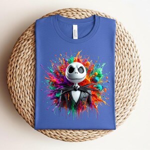 May include: A blue t-shirt featuring a graphic of a white skeletal character with a black pinstripe suit and bow tie, centered against a vibrant, multi-colored paint splatter background. The shirt has a white neck label with the text CANVAS.