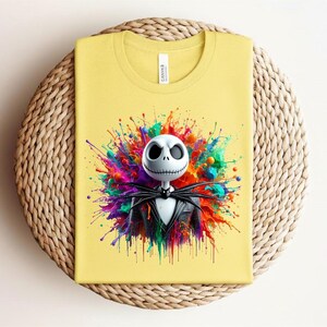 May include: A pale yellow t-shirt featuring a graphic print of a white skeletal character with large dark eye sockets and a stitched mouth, wearing a black pinstripe suit and bat-shaped bowtie, set against a vibrant, multi-colored paint splatter.