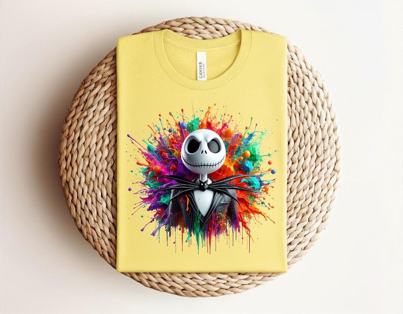 May include: A pale yellow t-shirt featuring a graphic print of a white skeletal character with large dark eye sockets and a stitched mouth, wearing a black pinstripe suit and bat-shaped bowtie, set against a vibrant, multi-colored paint splatter.