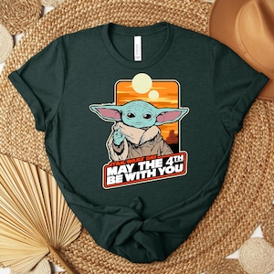 Może przedstawiać: Ciemnozielona koszulka z grafiką postaci z Gwiezdnych Wojen, z dużymi uszami i beżowym płaszczem. Grafika zawiera napis "STAR WARS DAY MAY THE 4TH BE WITH YOU" w kolorze białym i czerwonym. Koszulka jest złożona na tkanej powierzchni.