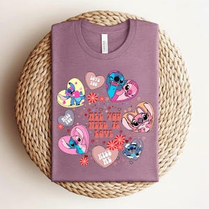 Peut inclure: Un t-shirt mauve avec un motif graphique color&eacute;. Le motif comprend des personnages de dessins anim&eacute;s en forme de c&oelig;ur, avec le texte "All You Need Is Love" et des phrases comme "Love You" et "Kiss Me".