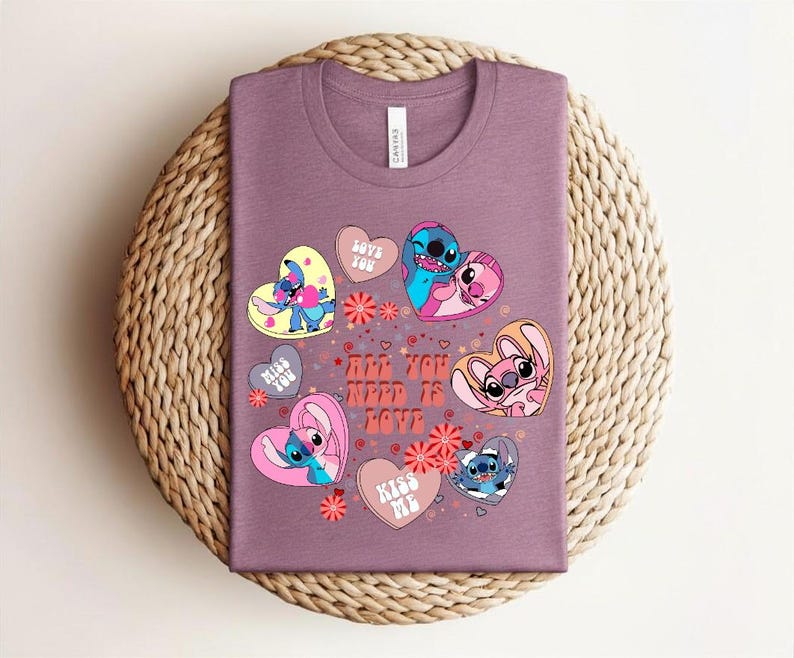 Peut inclure: Un t-shirt mauve avec un motif graphique color&eacute;. Le motif comprend des personnages de dessins anim&eacute;s en forme de c&oelig;ur, avec le texte "All You Need Is Love" et des phrases comme "Love You" et "Kiss Me".