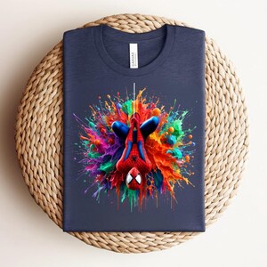 May include: Navy blue t-shirt featuring a colorful, artistic depiction of Spider-Man. The superhero is shown upside down, surrounded by a vibrant splash of paint in various colors. The shirt is folded on a woven, circular surface.