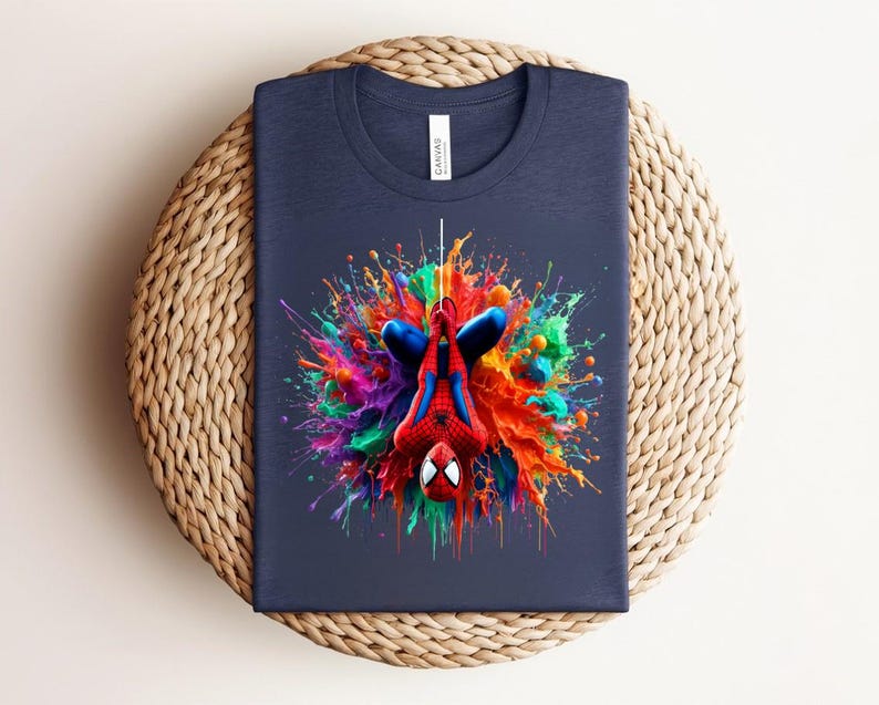 May include: Navy blue t-shirt featuring a colorful, artistic depiction of Spider-Man. The superhero is shown upside down, surrounded by a vibrant splash of paint in various colors. The shirt is folded on a woven, circular surface.
