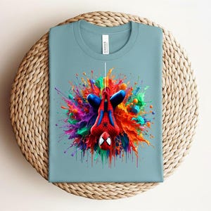 May include: A light blue t-shirt featuring a colorful graphic of Spider-Man hanging upside down. The superhero is surrounded by a vibrant explosion of paint in red, blue, yellow, and green. The shirt is folded on a woven surface.