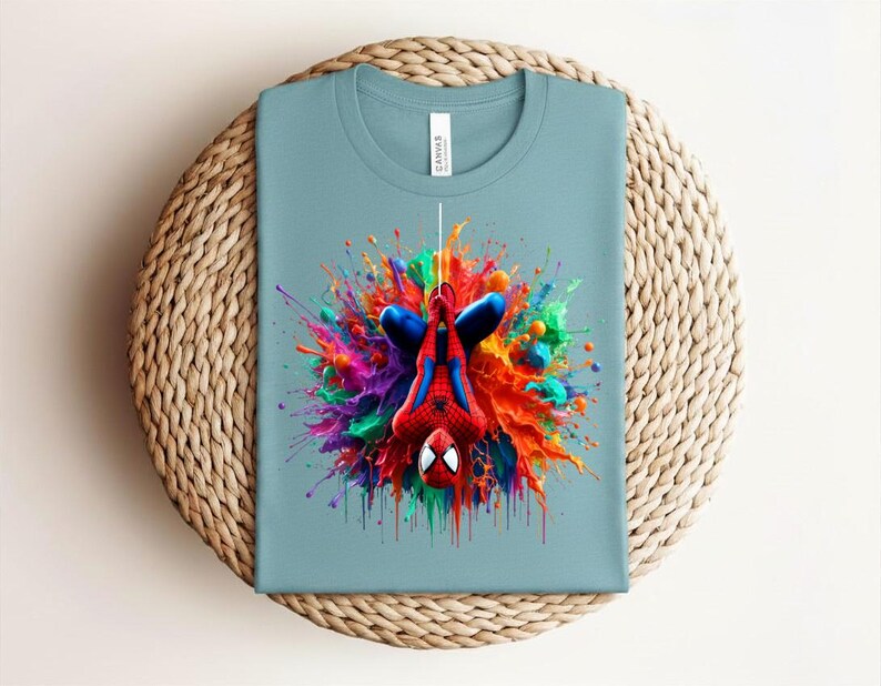 May include: A light blue t-shirt featuring a colorful graphic of Spider-Man hanging upside down. The superhero is surrounded by a vibrant explosion of paint in red, blue, yellow, and green. The shirt is folded on a woven surface.
