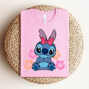 Puede incluir: Una camiseta rosa con un personaje de dibujos animados azul, ojos negros grandes y un lazo rosa. Flores de hibisco rosas y naranjas rodean al personaje. La camiseta est&aacute; doblada sobre una superficie circular tejida.