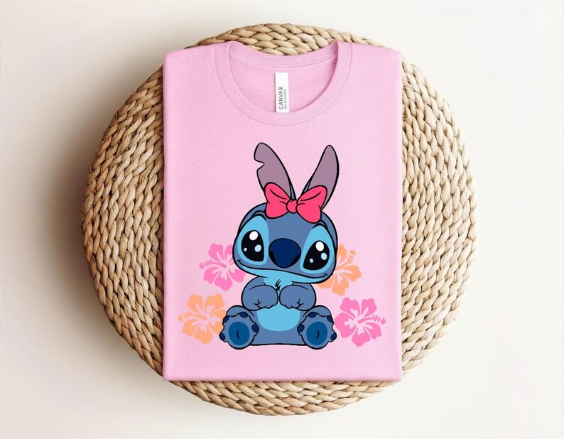 Puede incluir: Una camiseta rosa con un personaje de dibujos animados azul, ojos negros grandes y un lazo rosa. Flores de hibisco rosas y naranjas rodean al personaje. La camiseta est&aacute; doblada sobre una superficie circular tejida.